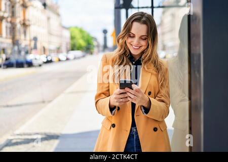 Femme souriante, messagerie texte via smartphone en attendant station de tram Banque D'Images