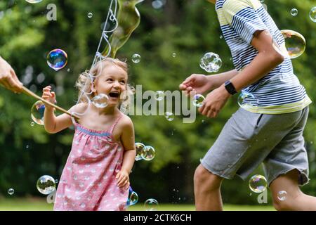 Fille joyeuse appréciant avec frère tout en courant au milieu des bulles à parc public Banque D'Images