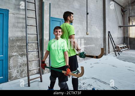 Garçon souriant tenant un bâton de hockey tout en se tenant dos à dos avec le père au tribunal Banque D'Images