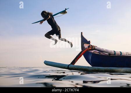 Homme avec planche de surf sautant en mer contre ciel bleu clair Banque D'Images