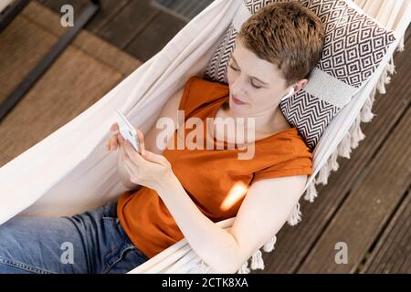 Femme aux cheveux courts utilisant un téléphone portable tout en se relaxant hamac dans le porche Banque D'Images