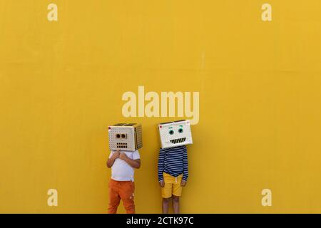 Garçons portant des masques robotisés faits de boîtes tout en se tenant contre mur jaune Banque D'Images