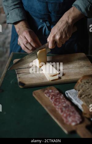 Section médiane du jeune homme coupant des tranches de fromage artisanales à bord à la table Banque D'Images