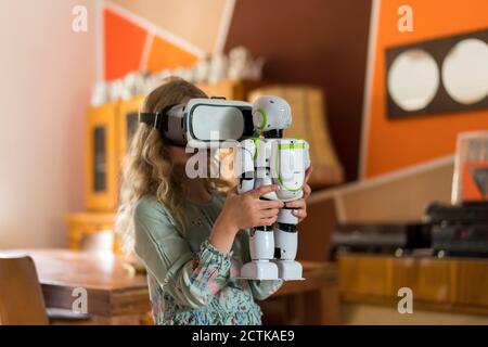 Fille avec un casque de simulateur de réalité virtuelle tenant le robot tout en jouant dans le salon à la maison Banque D'Images