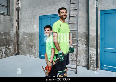 Garçon et père souriant tenant des bâtons de hockey tout en se tenant debout de retour sur le terrain Banque D'Images