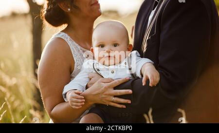 Section médiane des parents transportant un bébé garçon debout pendant le mariage à coucher de soleil Banque D'Images