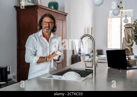 Homme souriant nettoyant la vaisselle tout en se tenant près de l'évier dans la cuisine Banque D'Images