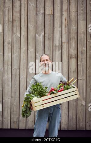 Homme mûr souriant portant une caisse de légumes tout en se tenant contre le mur Banque D'Images