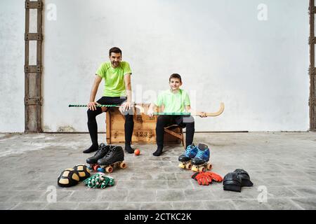 Père et fils assis avec des bâtons de hockey sur une caisse en bois par des équipements de sport contre le mur sur le terrain