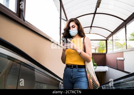 Jeune femme utilisant un téléphone portable debout avec un sac en filet sur la passerelle mobile du métro Banque D'Images