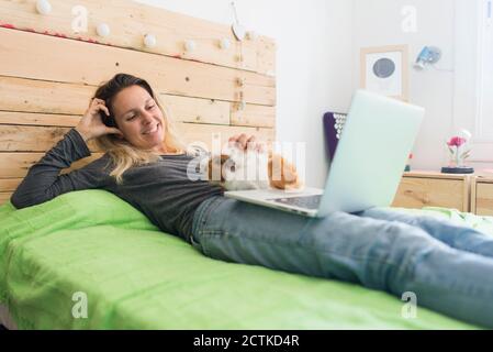 Une femme souriante qui a caressé un cobaye tout en étant allongée avec un ordinateur portable lit à la maison Banque D'Images