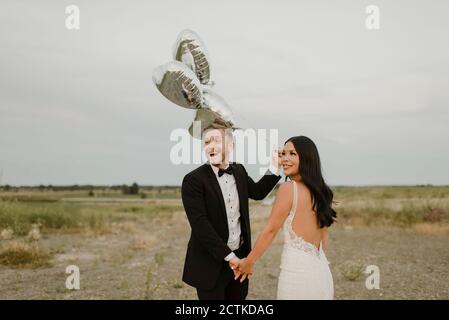 Joyeux marié avec des ballons en forme de coeur tenant la main de la mariée contre ciel Banque D'Images