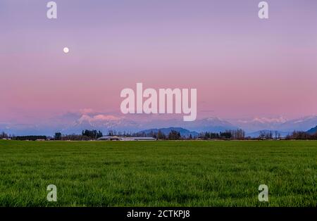 Magnifique paysage printanier, avec des montagnes enneigées en Colombie-Britannique, au Canada. Le coucher du soleil crée un magnifique ciel rose et pourpre. La pleine Lune brille b Banque D'Images