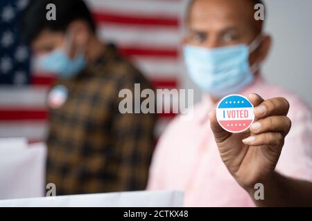 Gros plan des mains, l'homme dans un masque médical montrant que j'ai voté Sticker au kiosque de vote avec le drapeau américain comme arrière-plan - concept en personne votant à l'élection américaine. Banque D'Images