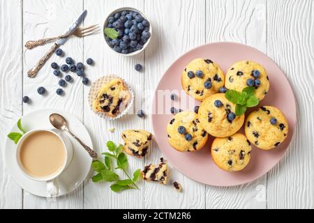 Muffins aux myrtilles cuits avec des myrtilles fraîches sur une assiette rose avec de la menthe fraîche sur le dessus et une tasse de thé avec du lait sur un fond en bois blanc, vue du dessus, Banque D'Images