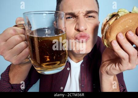 homme gai ivre avec une tasse de bière et un hamburger à la main régime alimentaire style de vie fond bleu Banque D'Images