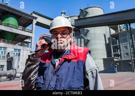 Mynaral/Kazakhstan: Ingénieur (travailleur) en casque blanc et lunettes de radio parlant. Silos et bâtiments industriels gris brouillé de l'usine de ciment Jambyl. Banque D'Images