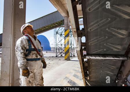 Usine de ciment Jambyl. Ingénieur ou technicien de maintenance en casque blanc et combinaison de protection blanche sale avec convoyeur à courroie en caoutchouc sur un support industriel Banque D'Images