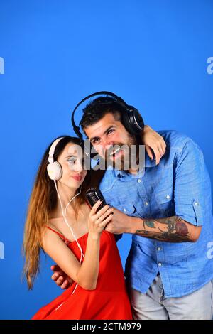 Concept de fête et de musique. Couple amoureux porte un casque et tient le microphone. Les fans de musique aux visages souriants aiment la musique et chanter. Homme avec barbe et fille accolade sur fond bleu. Banque D'Images