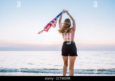 Vue arrière d'une jeune femme méconnue portant un drapeau américain au bord de la côte contre le coucher du soleil Banque D'Images