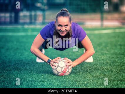 Bonne jeune athlète féminine souriante avec ballon de football dans le pose de planche de yoga avec vue frontale basse angle d'elle sourire à la caméra dans un cadre de santé et de fi Banque D'Images