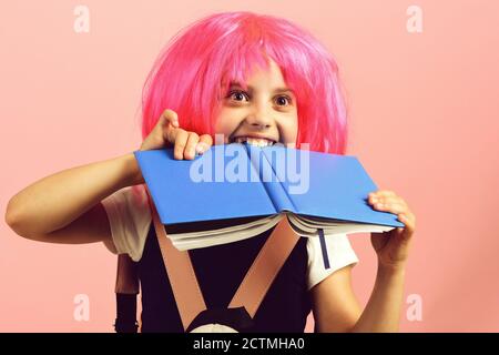 Élève en uniforme scolaire avec perruque rose. Fille morde grand livre bleu. Retour au concept d'école et d'éducation. Fille de l'école avec vue heureuse isolée sur fond rose. Banque D'Images