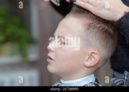 Coiffure fait une coiffure élégante. La femme est debout et fait la coupe de cheveux pour garçon blond. Vue rapprochée latérale. Banque D'Images