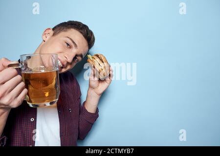 homme gai ivre avec une tasse de bière et un hamburger à la main régime alimentaire style de vie fond bleu Banque D'Images