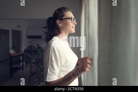 Femme heureuse avec des lunettes debout à côté d'une fenêtre et des rideaux d'ouverture à la maison, concept de style de vie Banque D'Images