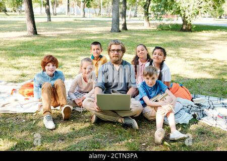 Portrait d'un jeune enseignant barbu en lunettes à l'aide d'un ordinateur portable avec groupe d'élèves du primaire dans le parc Banque D'Images