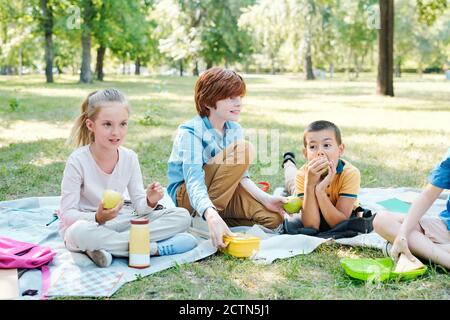 Groupe d'enfants assis sur des couvertures et en train de manger pendant le déjeuner pique-nique dans le parc après l'école Banque D'Images