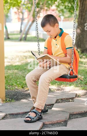 Smart boy avec sacoche orange assis sur balançoire et lecture réservez dans le parc après l'école Banque D'Images