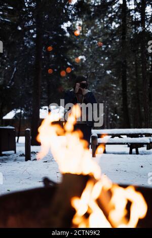 Explorateur méconnaissable dans des vêtements chauds prenant la photo de feu de joie brûlant avec des sparkles tout en se tenant sur le camping en forêt d'hiver pendant Voyager dans la nature aux États-Unis Banque D'Images