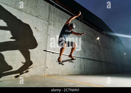 Angle bas de patineuse masculine talentueuse sautant avec la planche à roulettes tout en faire des cascades et s'entraîner dans un parc de skate éclairé la nuit Banque D'Images