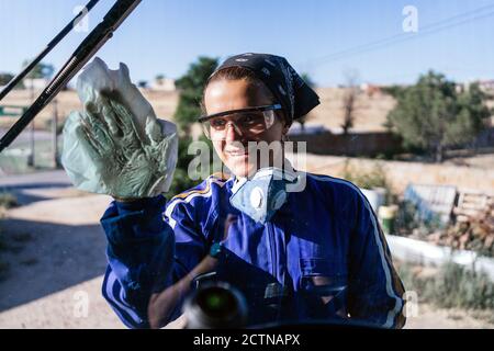 Femme souriante travaillant en uniforme et lunettes de protection pour nettoyer la fenêtre de la machine dans la zone industrielle Banque D'Images