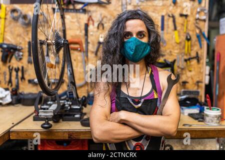 Femme mécanicien de souche adulte sérieuse en vêtements de travail et masque de protection tenez la clé et regardez la caméra tout en vous tenant contre les intempéries établi avec divers outils dans un atelier d'entretien de vélo Banque D'Images