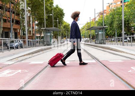 Vue latérale de l'homme africain avec cheveux afro et un masquer la marche avec une valise sur les roues lors de la traversée d'un tramway dans une ville Banque D'Images