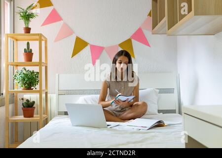 Femme étudiante assise sur le lit avec ordinateur portable et réserver pendant écrire dans un carnet et préparer l'examen à la maison Banque D'Images
