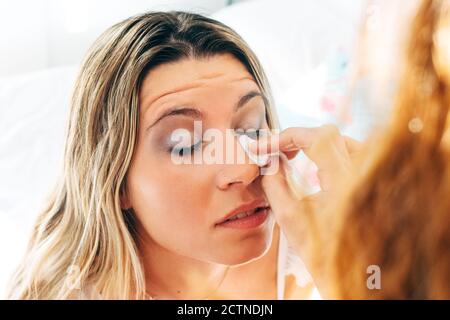 Femme écourtée non reconnaissable faisant le maquillage pour une amie avec les yeux fermés assis sur le lit à la maison Banque D'Images