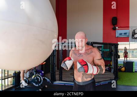 Déterminé boxer mâle dans des gants de boxe poinçant le sac lourd pendant entraînement intense dans la salle de gym Banque D'Images