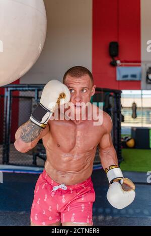 Déterminé boxer mâle dans des gants de boxe poinçant le sac lourd pendant entraînement intense dans la salle de gym Banque D'Images