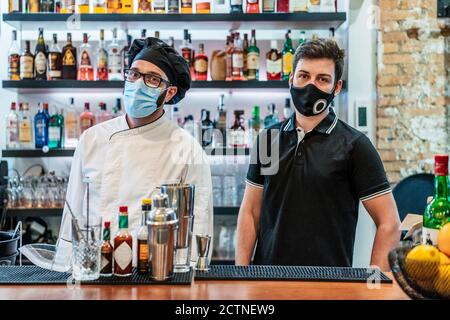 Cuisinier et barman sérieux dans des masques médicaux debout compteur dans la barre et regarder la caméra pendant l'épidémie de coronavirus Banque D'Images