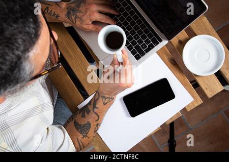 De dessus de la récolte mâle non reconnaissable dans les vêtements décontractés avec bras tatoués buvant du café et parcourant un ordinateur portable tout en travaillant à distance à la table dans le lieu de travail à domicile Banque D'Images