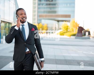 Ravi homme d'affaires afro-américain en costume formel marchant avec ordinateur portable dans la rue et conversation sur smartphone tout en discutant projet d'entreprise et regarder loin Banque D'Images