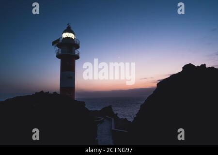 Faible angle de paysage spectaculaire de phare éclairé situé sur colline des rocheuses près de la mer contre le ciel de coucher de soleil à Ténérife Banque D'Images