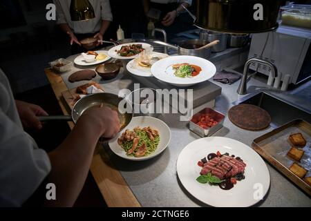Grand angle de récolte anonyme chef ajoutant des filets de poulet frits dans l'assiette avec des pâtes tout en préparant des aliments avec des collègues cuisine moderne Banque D'Images
