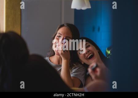 Des amies ravies debout devant le miroir à la maison et faire le maquillage ensemble tout en préparant la fête Banque D'Images