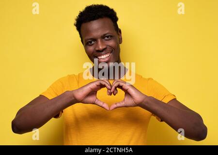 Beau sourire jeune homme africain fait le coeur en utilisant les doigts sur le mur jaune. Banque D'Images