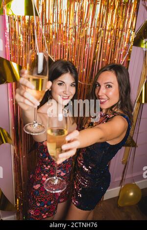 Jeunes amies gaies dans des robes de fête stylées qui applaudissent Des verres de champagne et regarder l'appareil photo tout en appréciant New Fête de l'année Banque D'Images