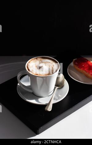 Tasse en céramique blanche servie avec du café frais recouvert de lait faire mousser et faire du pain avec de la confiture Banque D'Images